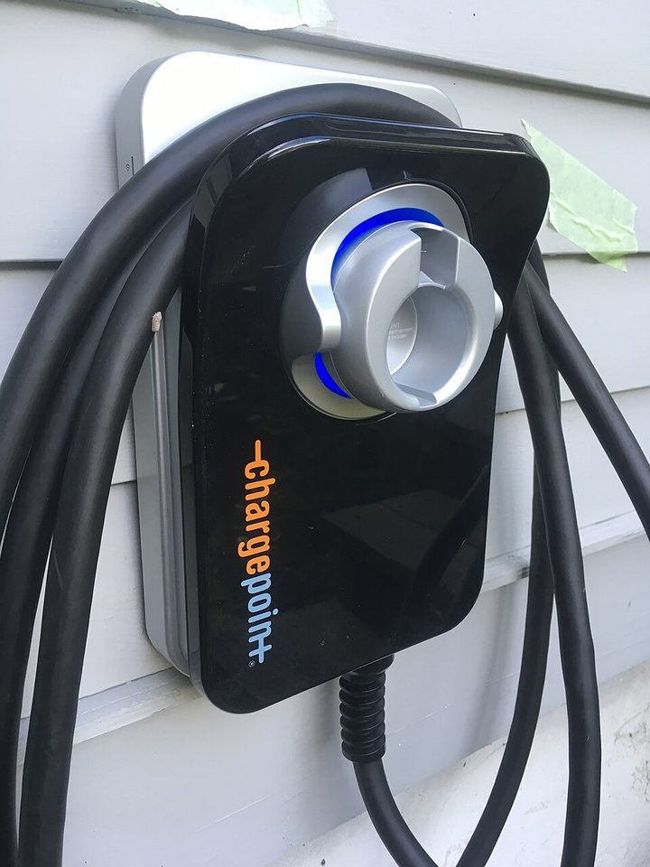 A home electric vehicle charger mounted on the lap siding of a house, its connector holstered in the face of the unit and the cable looped to one side.