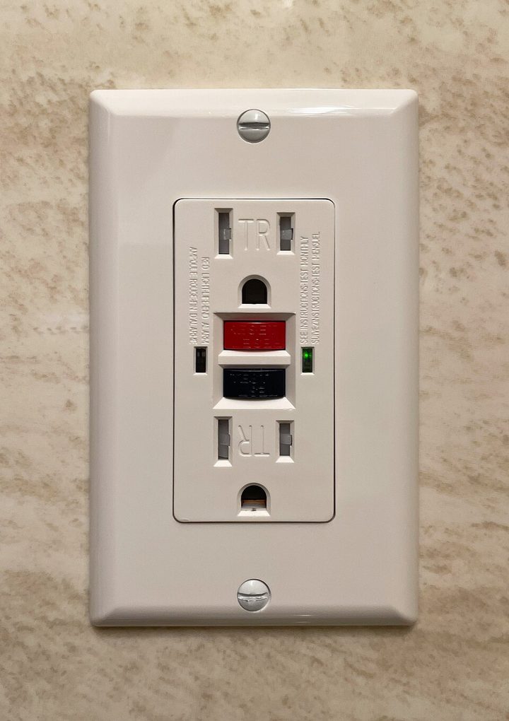 A white GFCI receptacle in a wall plate, with TEST and RESET buttons between its two outlets and a small green status light beside them.