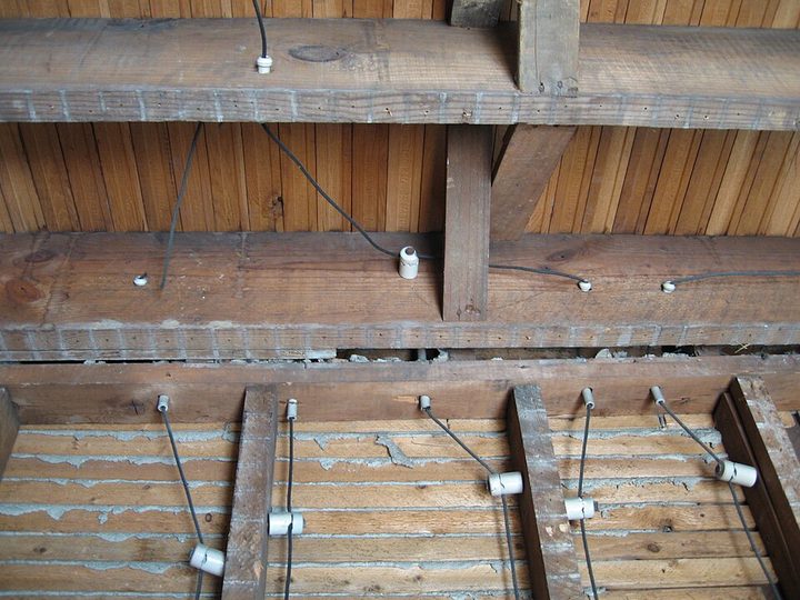 Knob-and-tube wiring above a lath ceiling: single conductors held on white ceramic knobs and passed through ceramic tubes where they cross wooden joists.