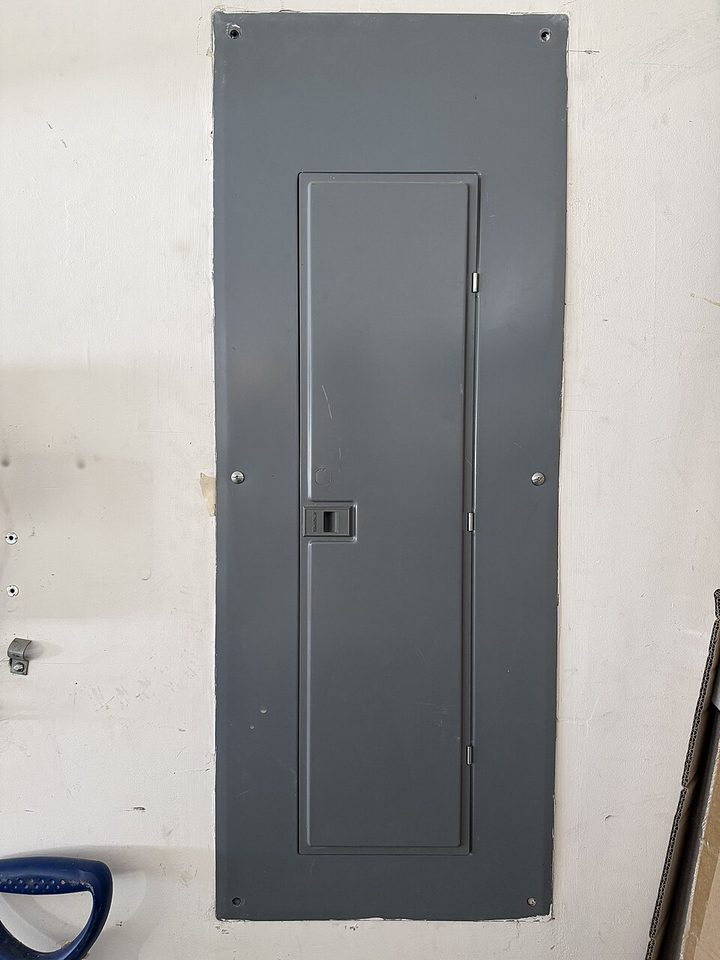 A gray flush-mounted breaker panel set into a painted basement wall, its door closed.