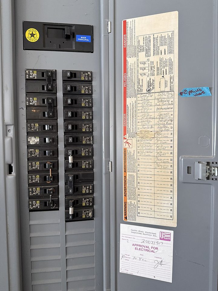 A residential breaker panel with its door open. The main breaker is at the top, a hand-written circuit directory lists rooms and appliances beside it, and a signed electrical inspection approval sticker is fixed below that.