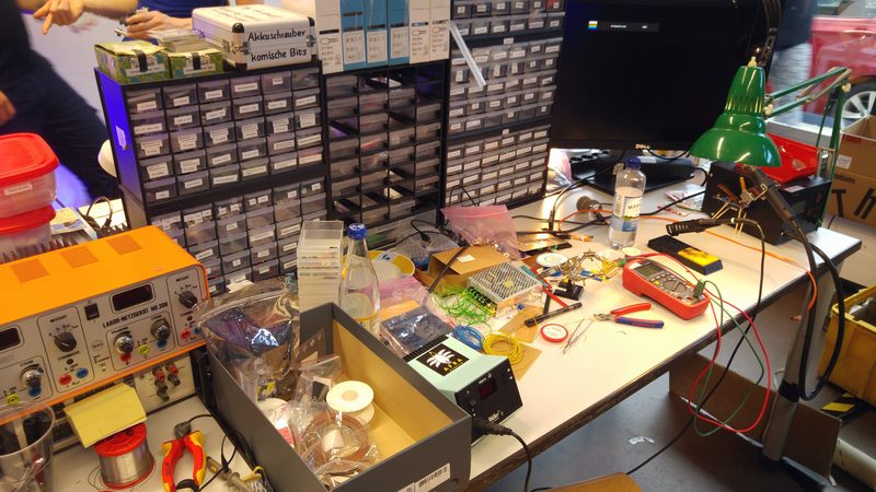 A repair bench along its length: a soldering station, a wall of small component drawers, spools of solder and hand tools under a lamp.