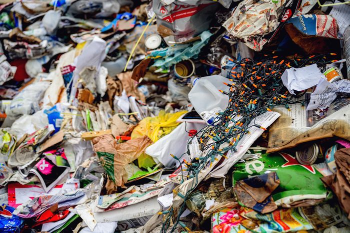 Close on a heap of mixed recyclables: cans, cartons, film plastic, paper and a tangle of string lights.