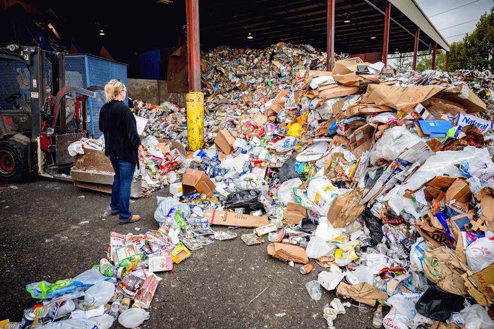 A worker seen from behind in a recycling sorting shed, a loader beside them and a mound of mixed material running away to the right.