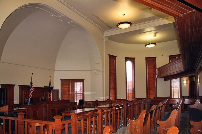 An empty historic county courtroom: rows of wooden pews, two flags beside the bench and tall windows down one side.