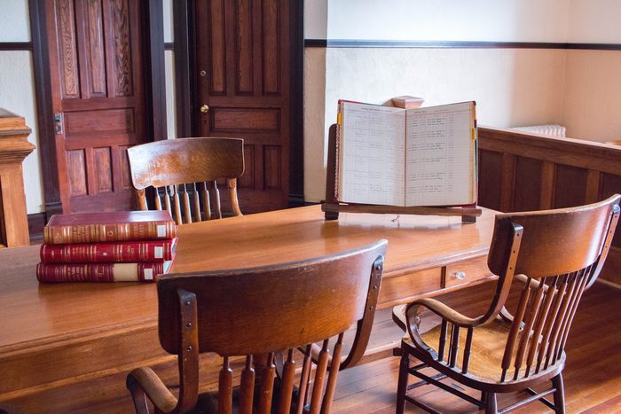 Close on a counsel table in a courthouse: an open ledger on a stand, a stack of bound law books and two spindle-back chairs.