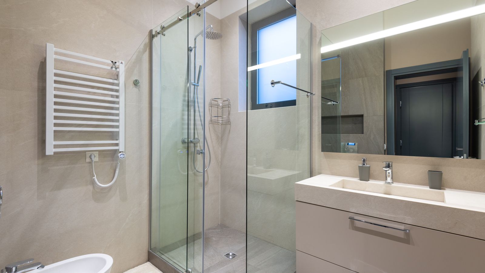 The finished bath: a glassed shower with a flush drain, a long stone vanity under a lit mirror, and a heated towel rail.