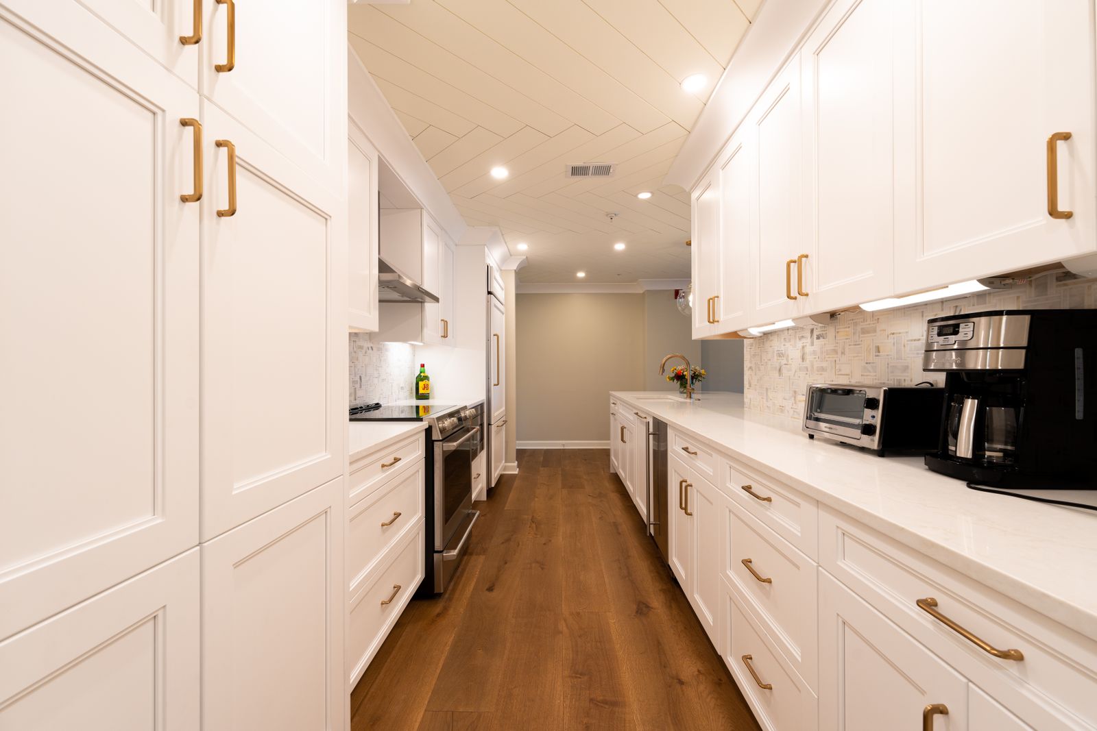 A galley kitchen with white cabinetry, brass hardware and a run of wood flooring.