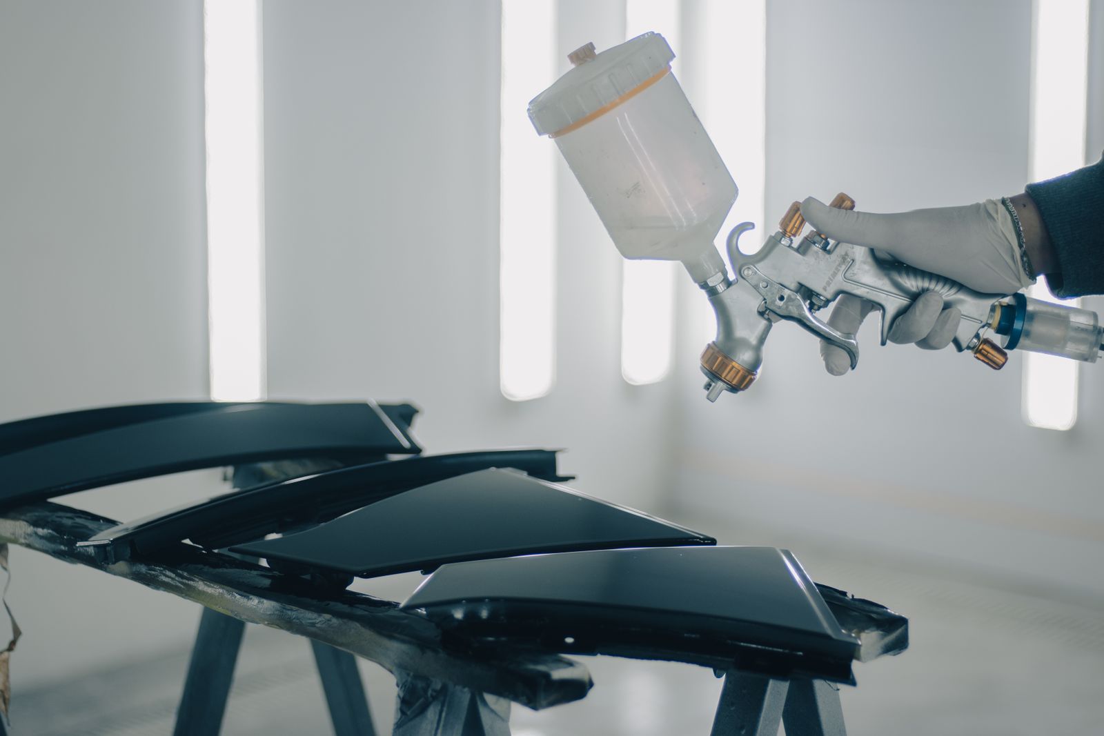 A gloved technician spraying detached vehicle panels inside a paint booth