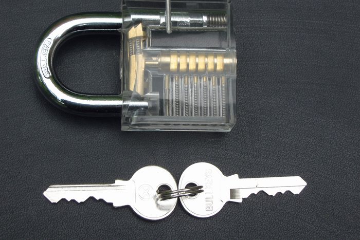 A cutaway padlock with a transparent body showing the pin stacks and springs inside it, two cut keys lying below.