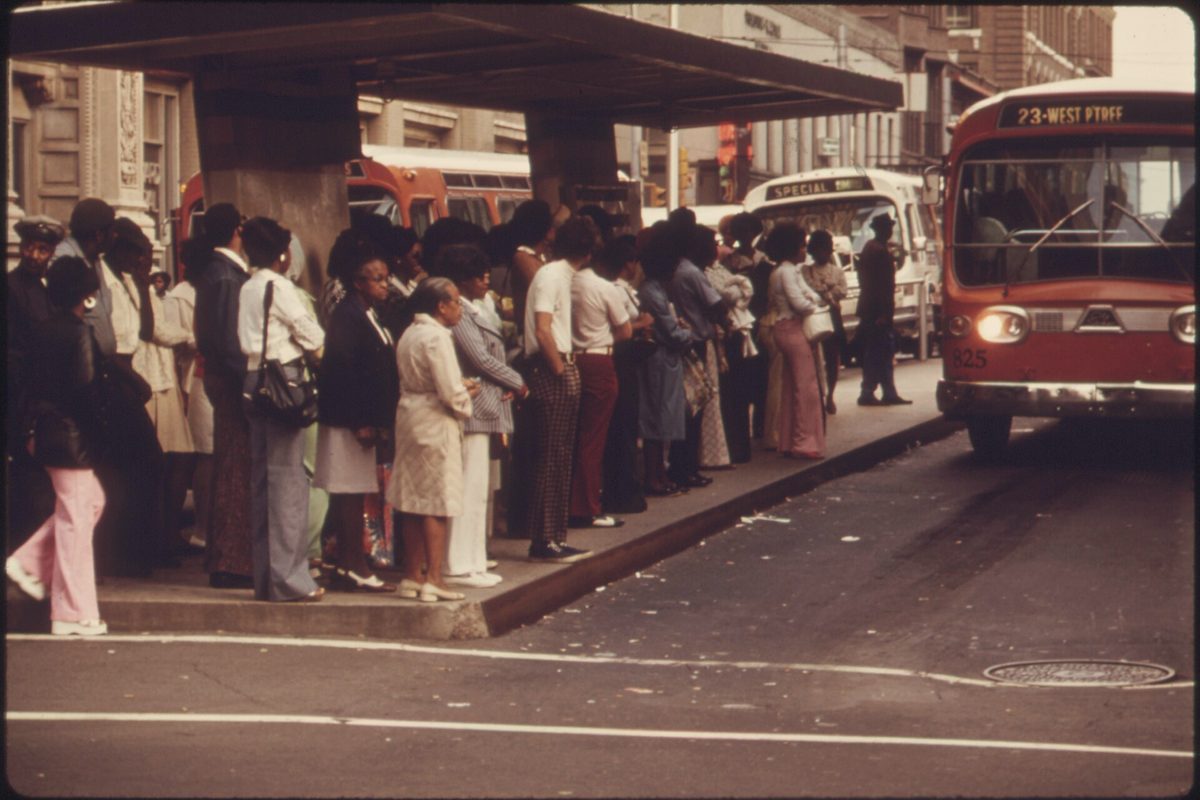 A queue of people waiting to board a city bus under a shelter, photographed in the 1970s