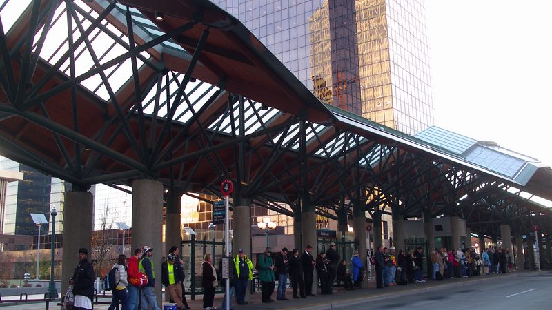 A transit center platform under a long curved canopy, a line of people waiting along the curb and towers behind.