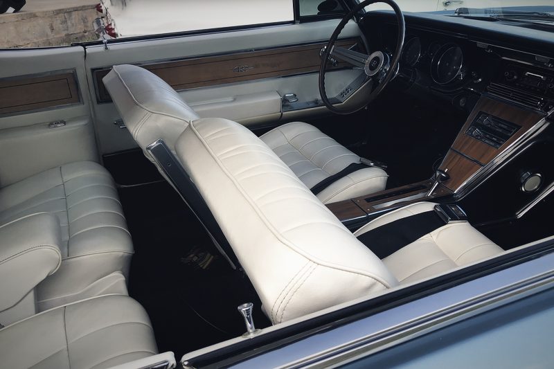 The white leather interior of a Buick Riviera, showing the wood-rimmed steering wheel and bench seat.
