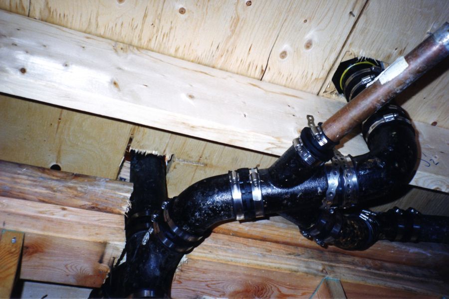 Black cast-iron drain, waste and vent piping running beside copper supply lines under a wood floor.