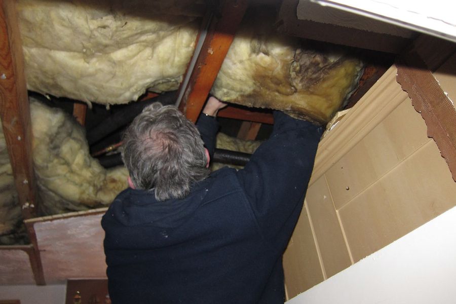 A plumber reaching up into an insulated joist bay to work on a pipe run.